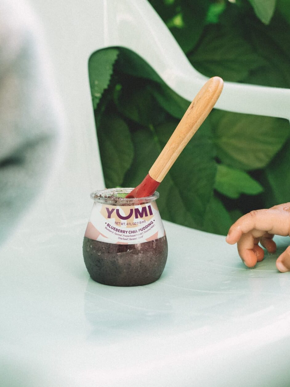 A jar of Yumi blueberry chia pudding sits on a chair with a wooden spoon.