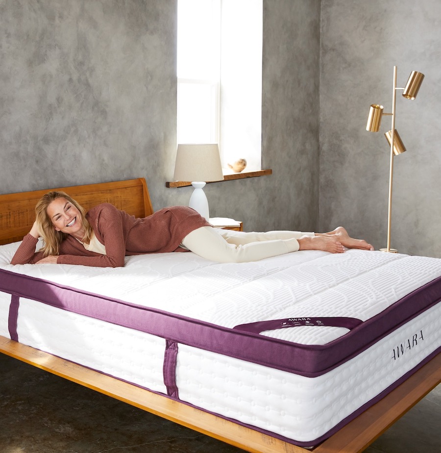 A bare Awara mattress which is white with purple trim on the edges fills a grey bedroom and a woman in her fifties lays out on top.