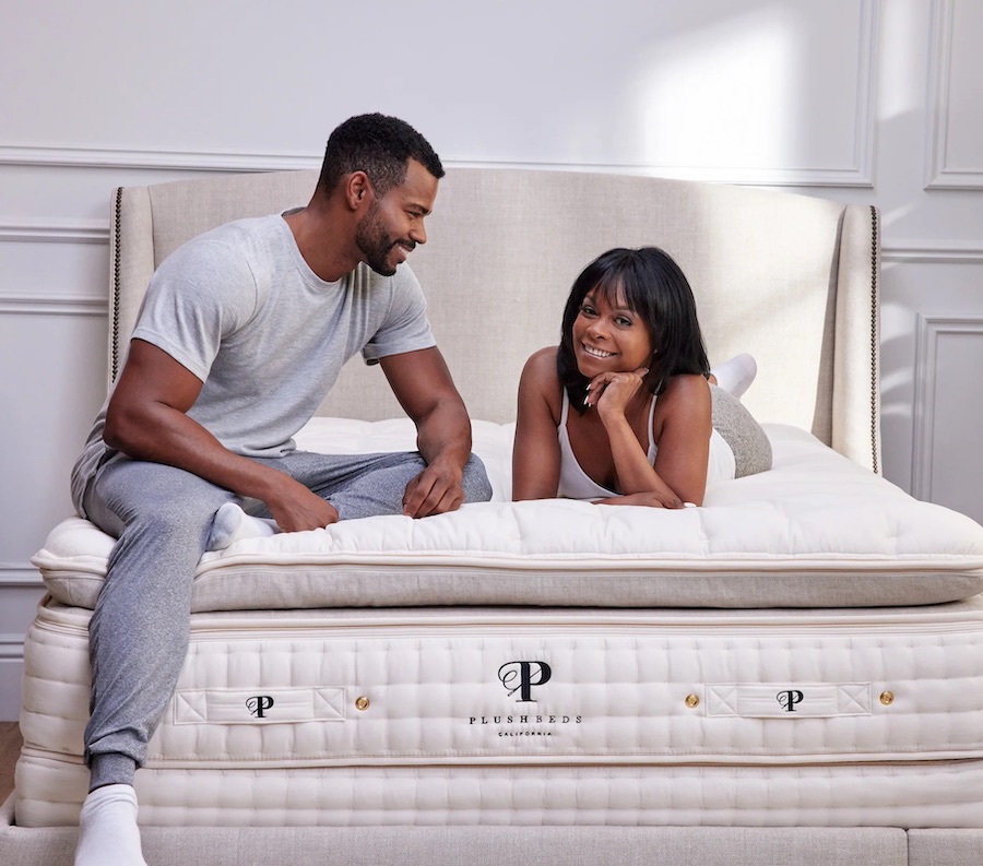 A couple lay on top of a very plush mattress from Plushbeds, there is no bedding on it so you can see the Plushbeds logo