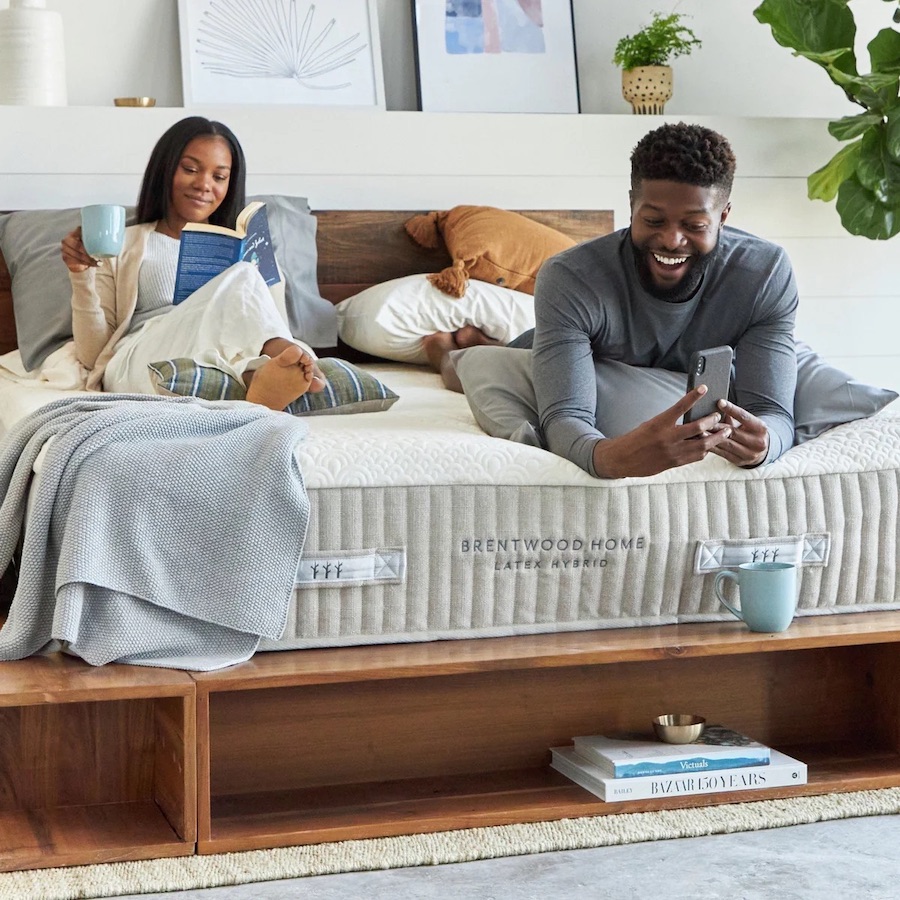 A couple reads on top of a Brentwood Home Latex Hybrid mattress that has no sheets so the logo is visible