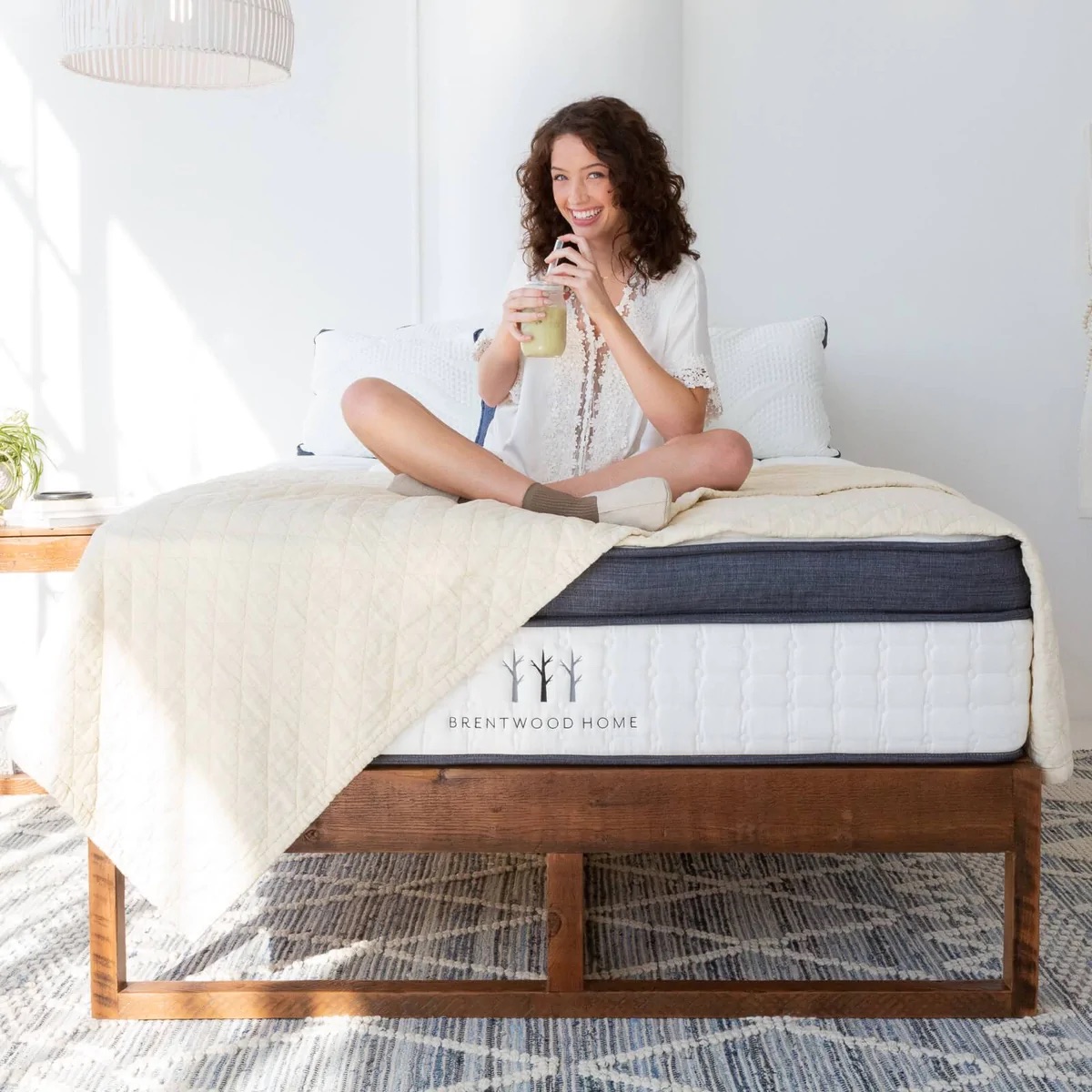 A woman smiles and drinks a smoothie while sitting on top of a mattress from Brentwood Home, the logo is clearly visible