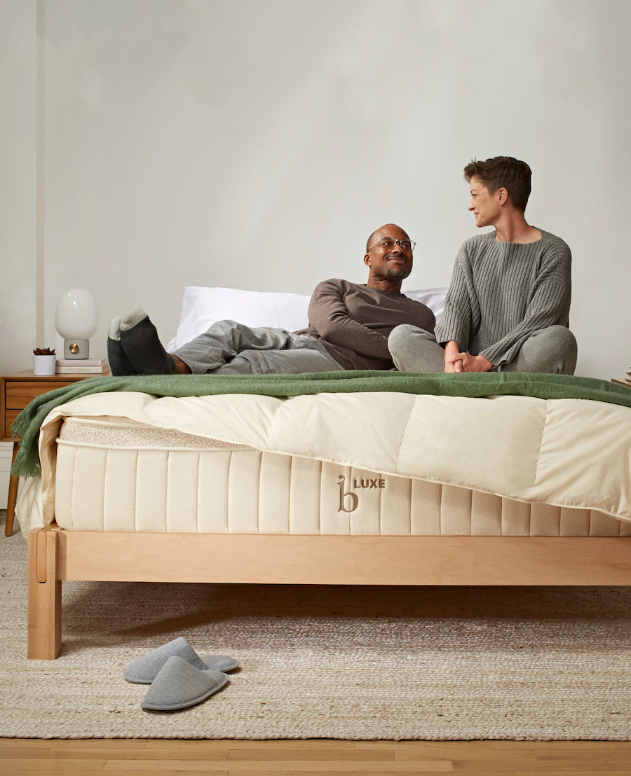 A couple in loungewear sit and lay on top of a green blanket that is draped across a Birch Luxe organic mattress