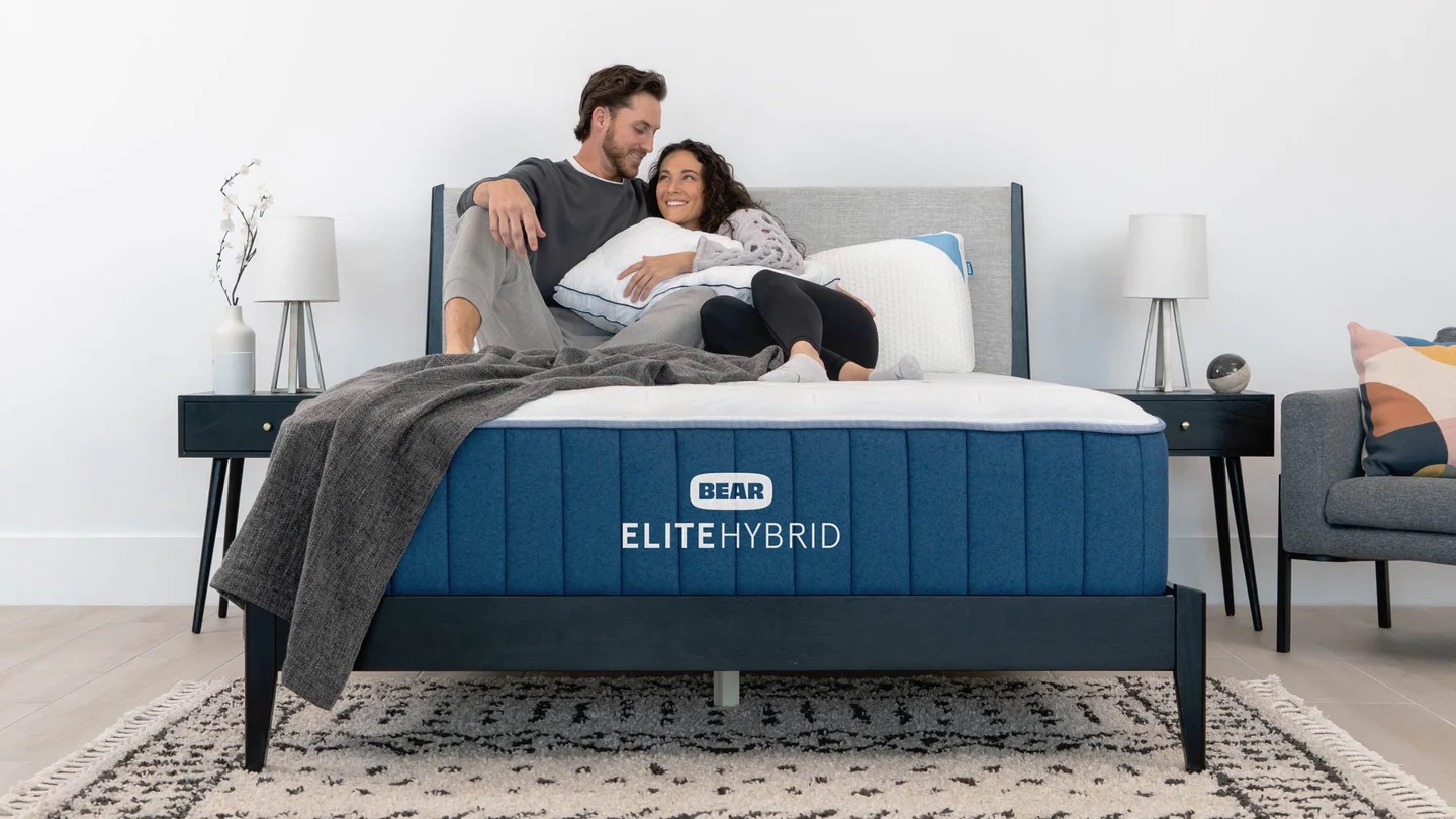 A couple snuggles on top of a mattress that does not have sheets on it, and you can see the Bear: Elite Hybrid logo at the foot of the bed