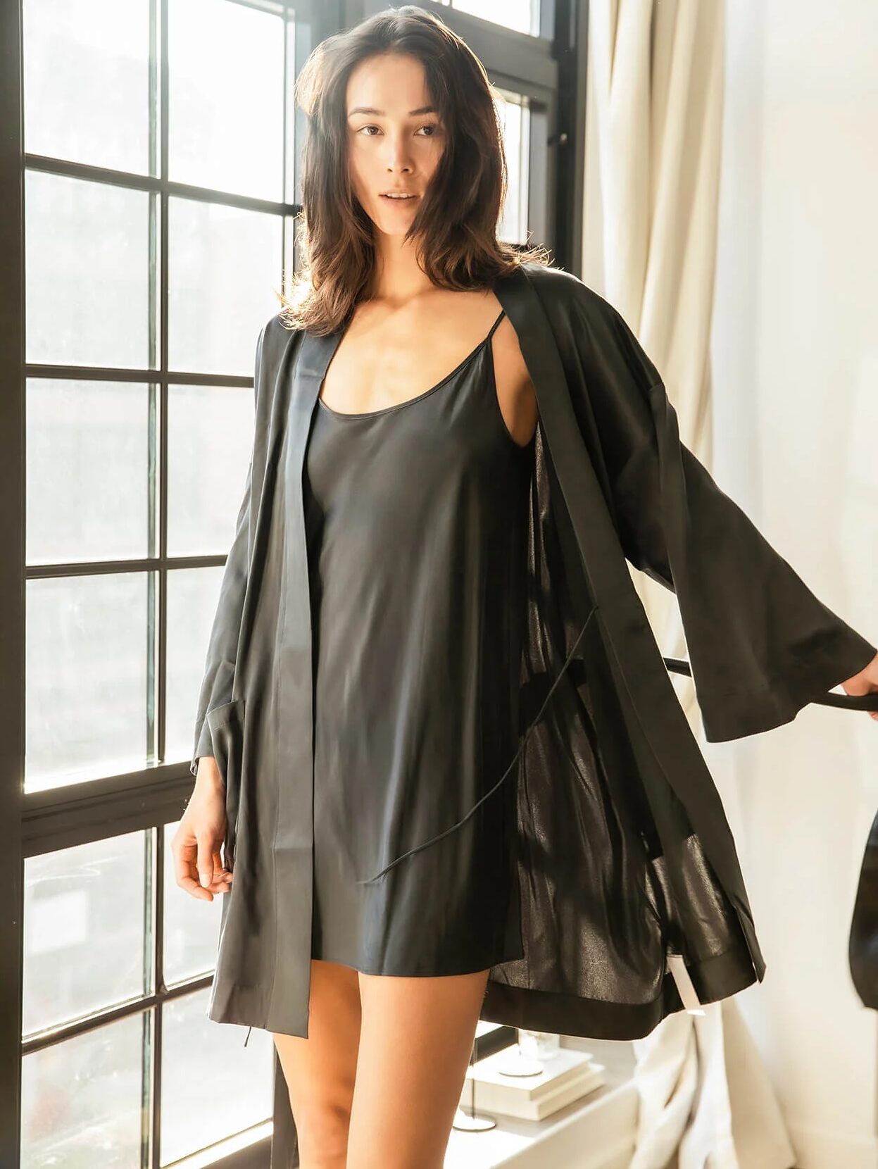 A model in a black silk robe and matching nightgown stands in front of a window and looks at the camera