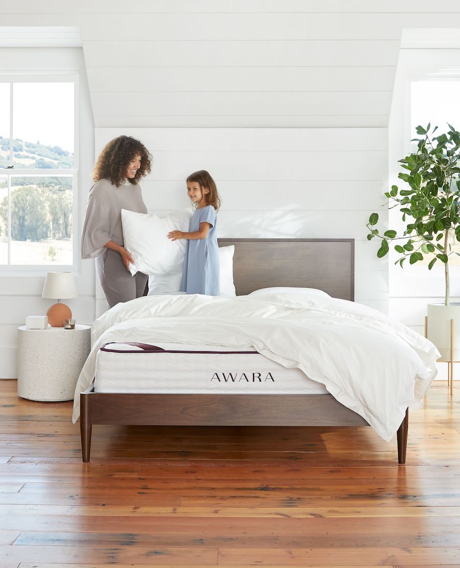 A woman stands by an Awara organic mattress that is topped with a fluffy comforter and hands a young child a fluffy pillow