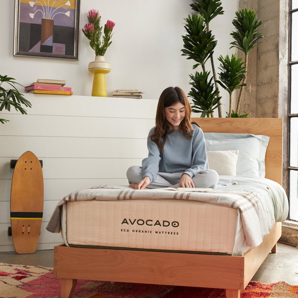 A young girl with brown hair sitting cross legged and reading on top of an Avocado Eco Organic Mattress in a twin size.