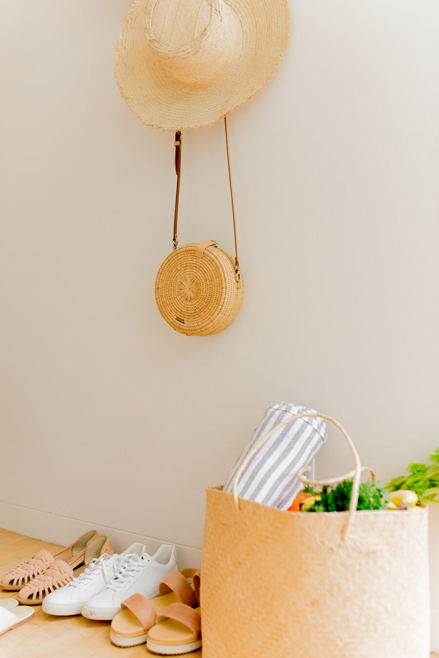A straw hat and round bag hang on a white wall above a row of shoes, with a woven tote bag filled with groceries and a striped towel on the floor.