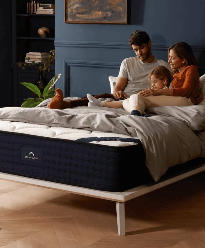 Two parents and a child snuggle on top of a bed reading together, the blankets are pulled up to reveal a dreamcloud logo.