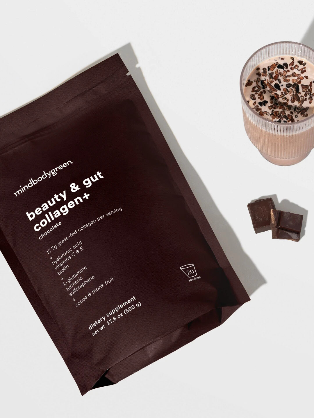 A pack of mindbodygreen chocolate collagen next to two piece of chocolate and a chocolate drink.