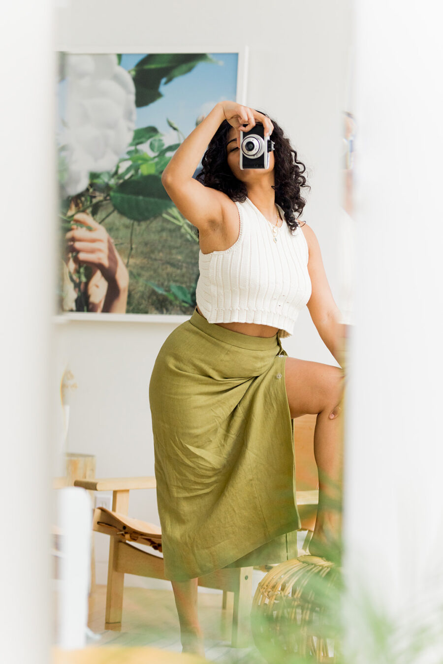 Woman in white sweater tank top and green linen skirt snaps a picture of herself in the mirror using a vintage film camera.