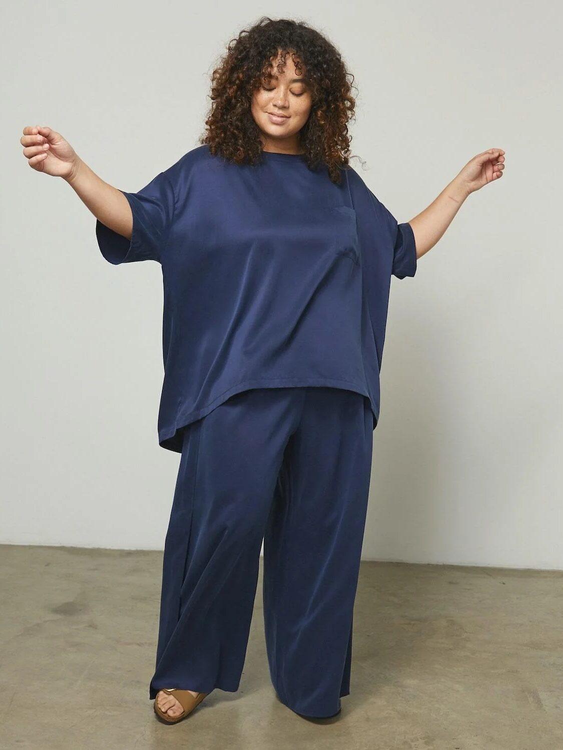 A model wearing a navy silk tee and wide leg pants set holds our her arms and smiles as she looks at the floor