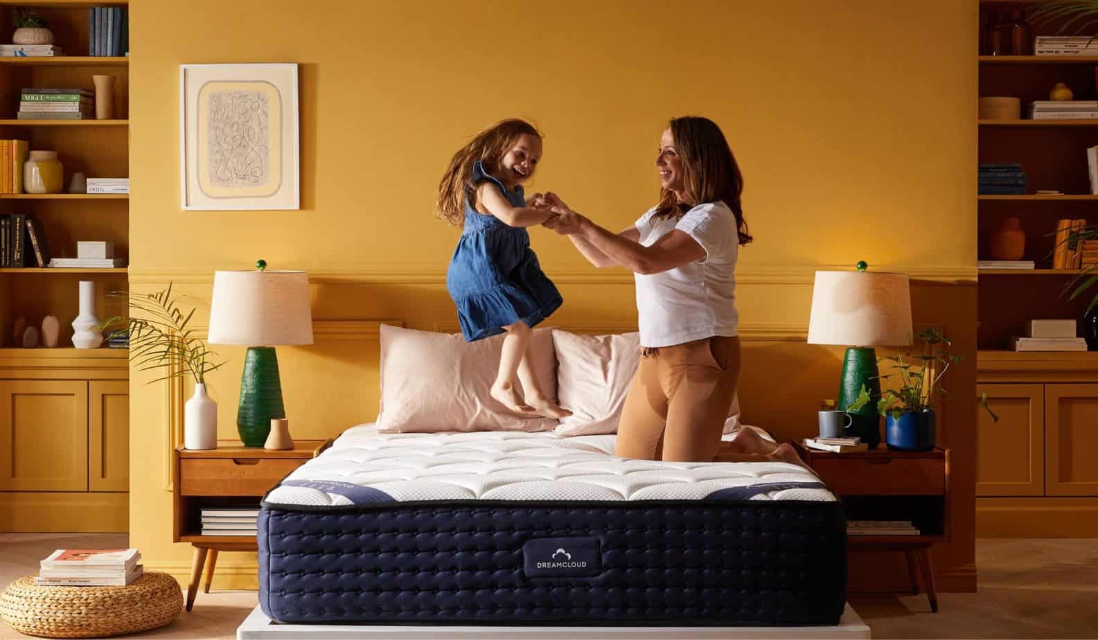 A child jumps on top of a bare dreamcloud mattress in a yellow room, while holding her mom's hands.