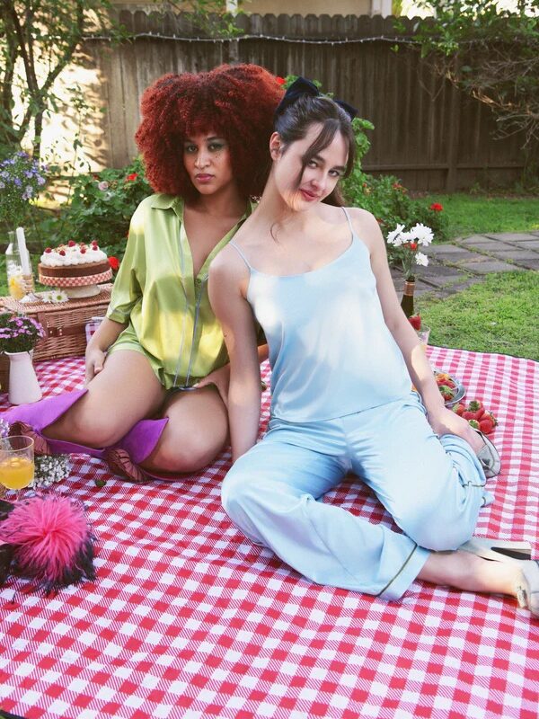 Two models sit on a red gingham picnic blanket wearing colorful silk pajamas