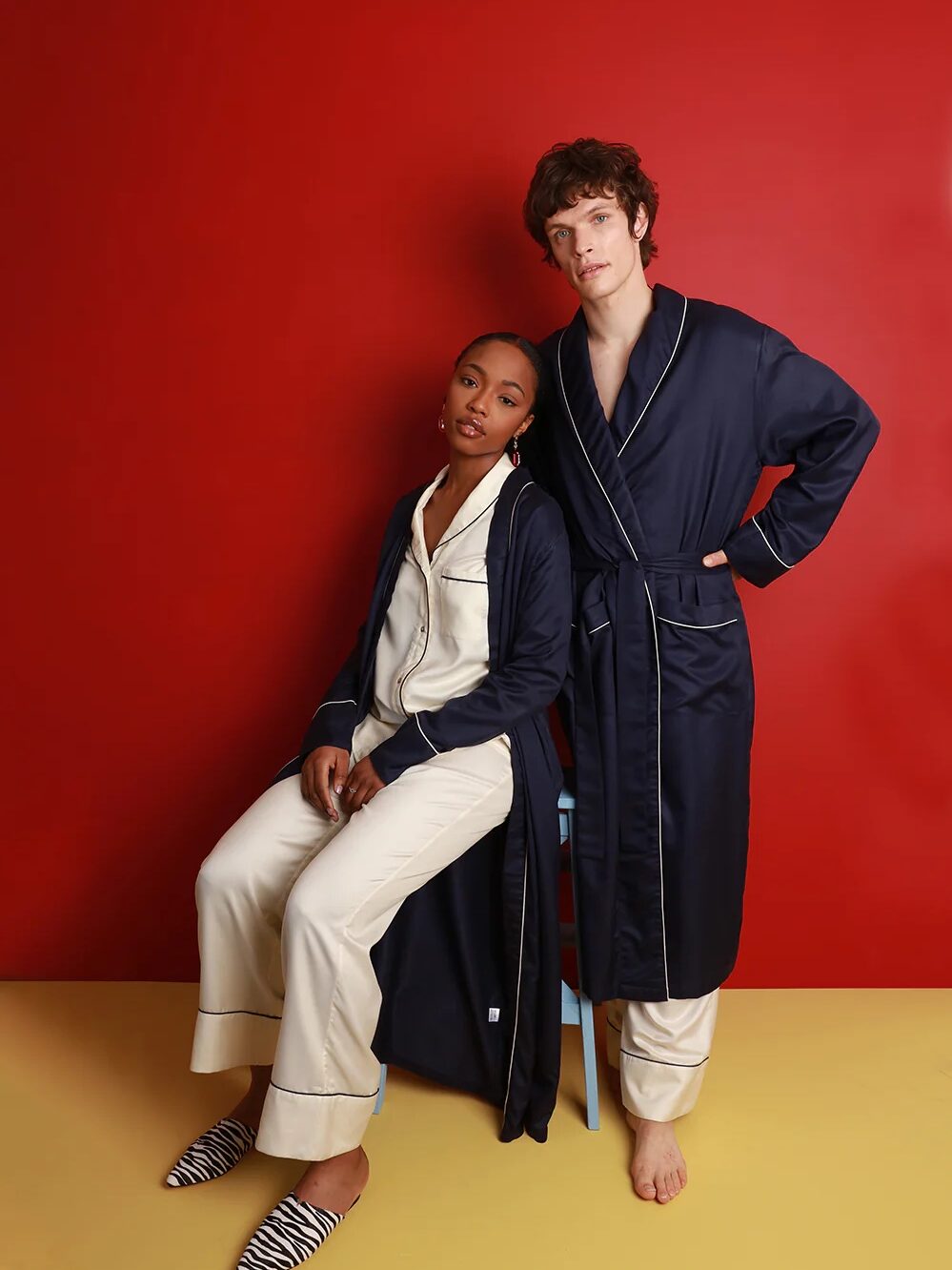 A woman seated in front of a man wearing matching white silk pajamas with black piping and navy silk robes with white piping look at the camera
