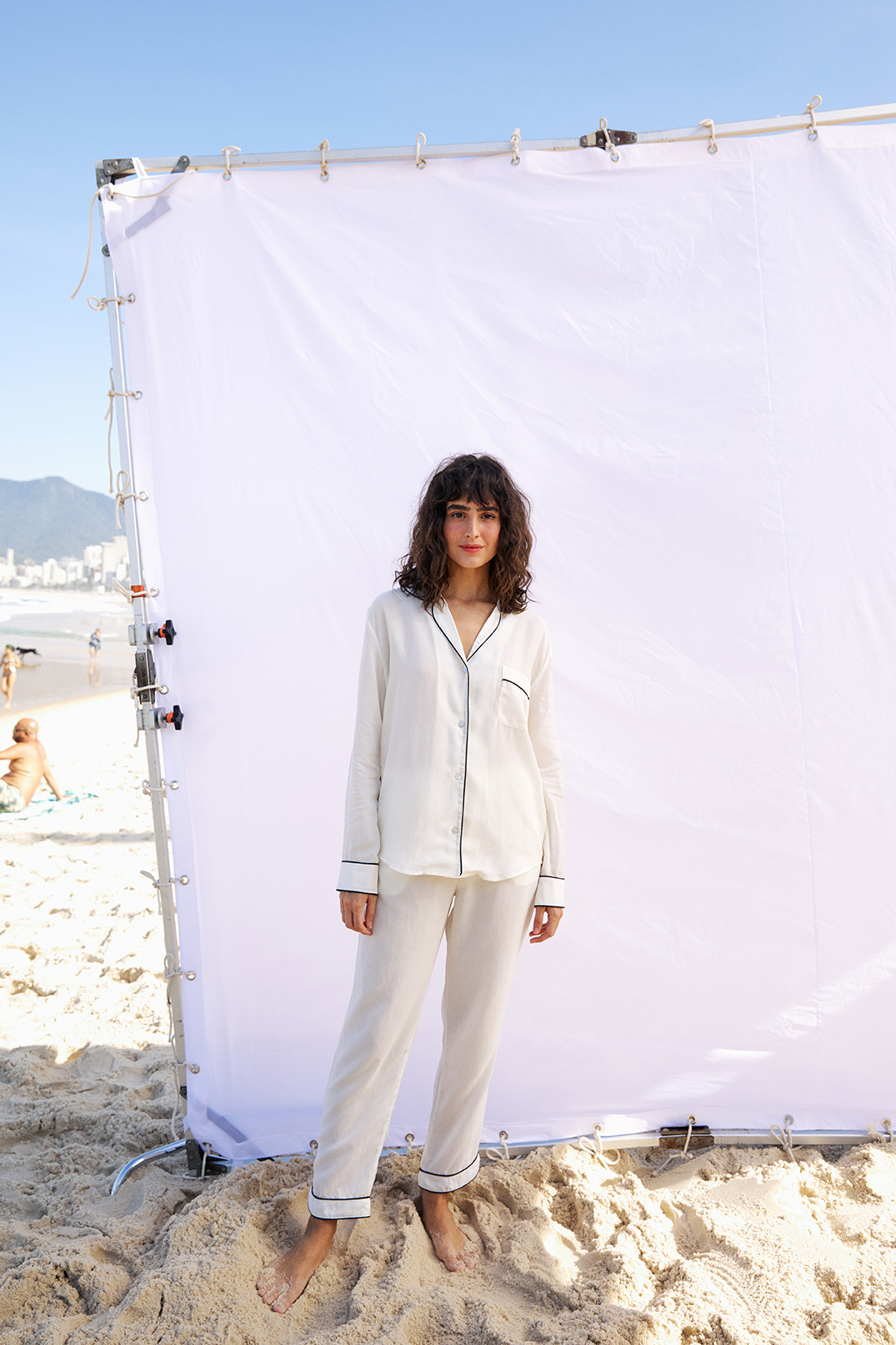 A model in front of a white photo backdrop on a beach wears  white silk pajamas with black piping