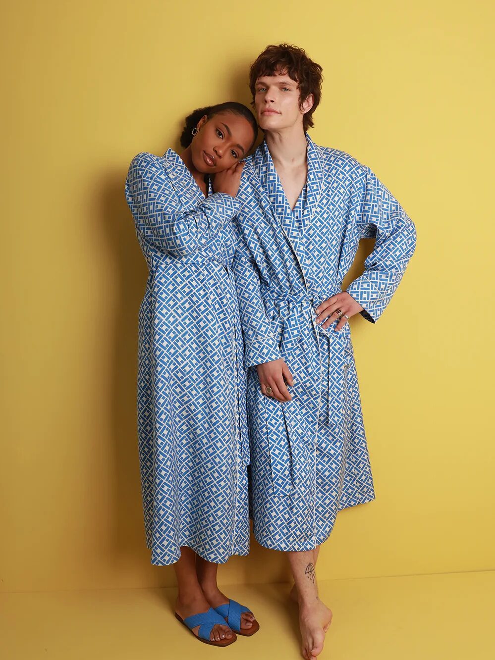 A man and a woman in matching blue print silk robes look at the camera and hold each other in front of a yellow wall