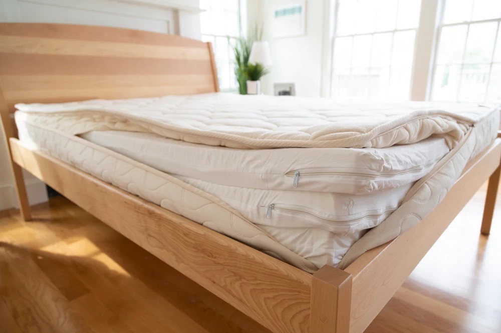 A bare mattress on a light wood bed frame is unzipped to reveal three separate layers of latex