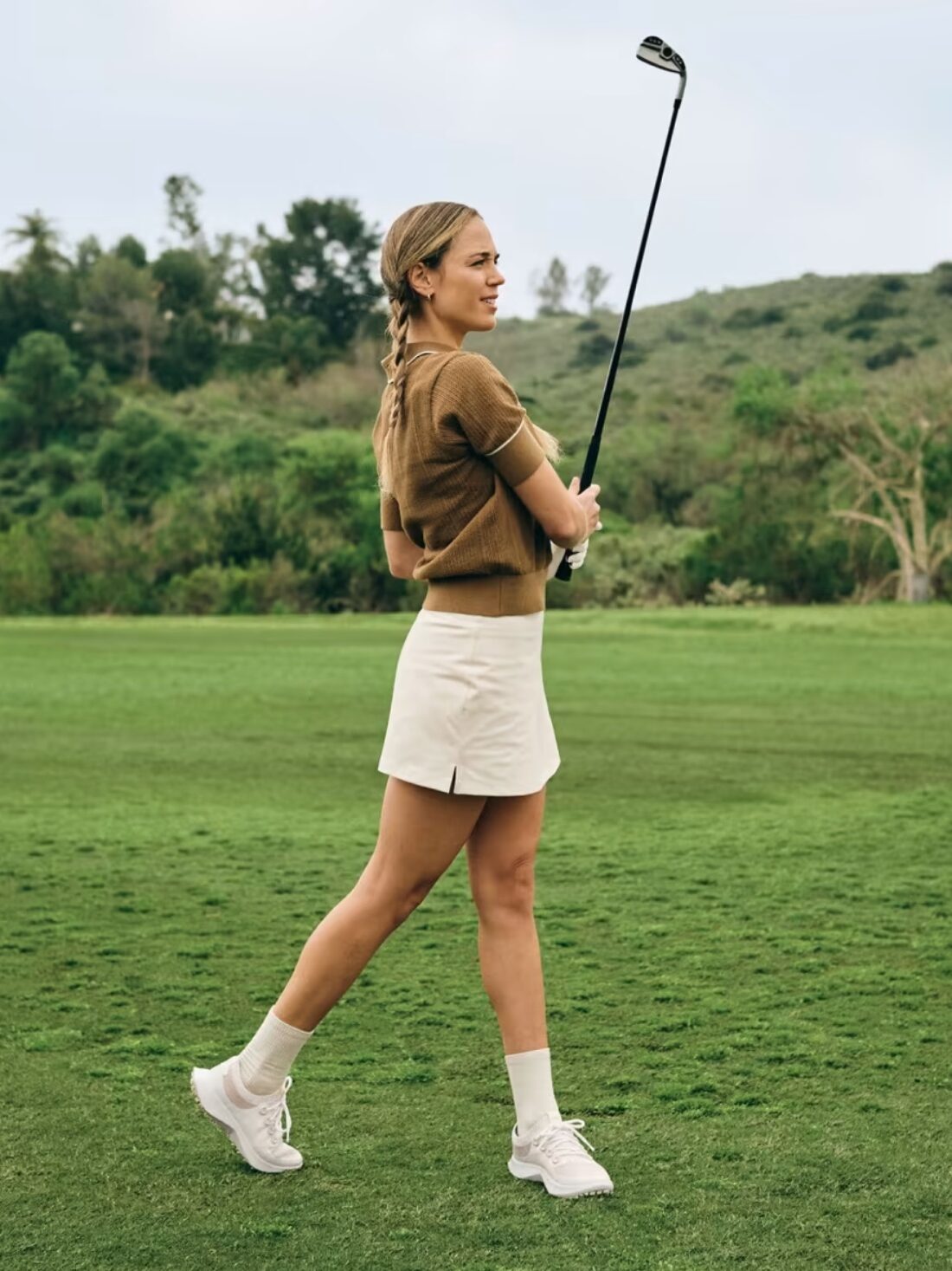 Model in white tennis shoes and skirt and brown shirt holds a golf club