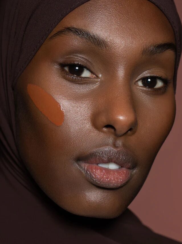 A close up of a Black model in a head scarf shows a swatch of the produce on her cheek.