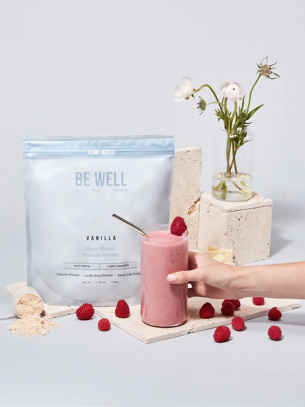 A bag of Be Well protein powder sits in a tableau with a pink smoothie, some scattered raspberries, a vase of white ranunculus flowers, and a few white stone blocks.