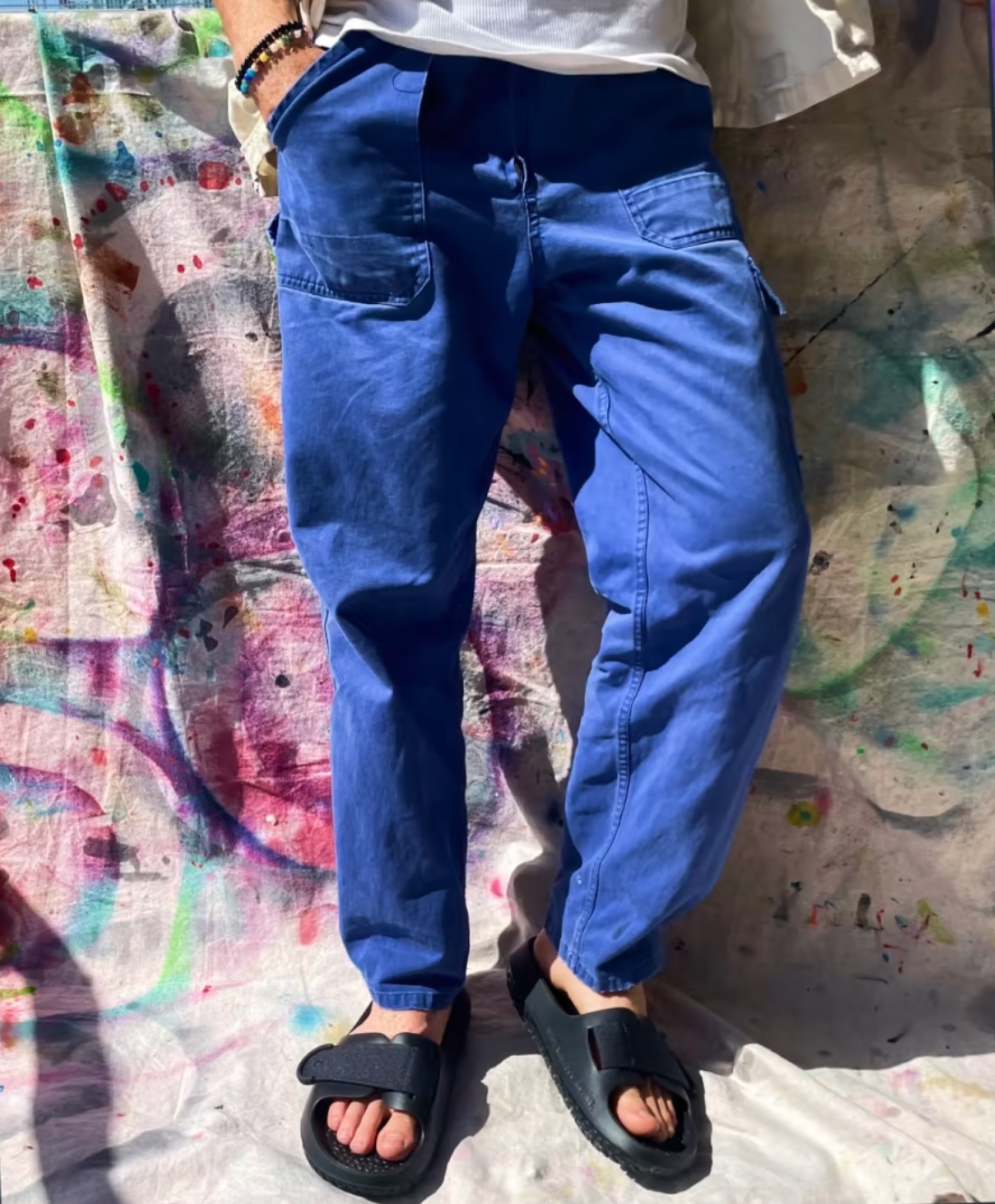 The lower half of a person's body leans against a painted canvas drop cloth, wearing blue pants and black Allbirds sugar glider sandals.