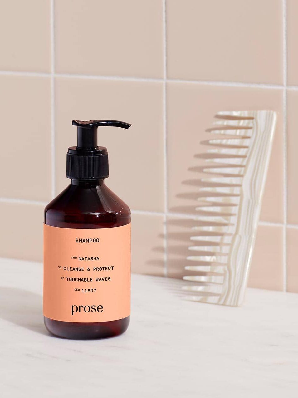 The shampoo product sits on a bathroom counter next to a comb propped against a pink tiled wall.
