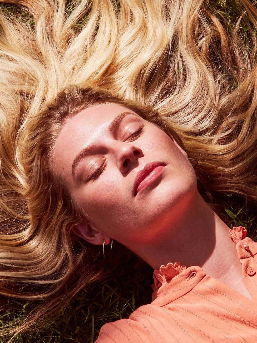 A model lies on the ground with her eyes closed and her long blond hair fanned around her head. 