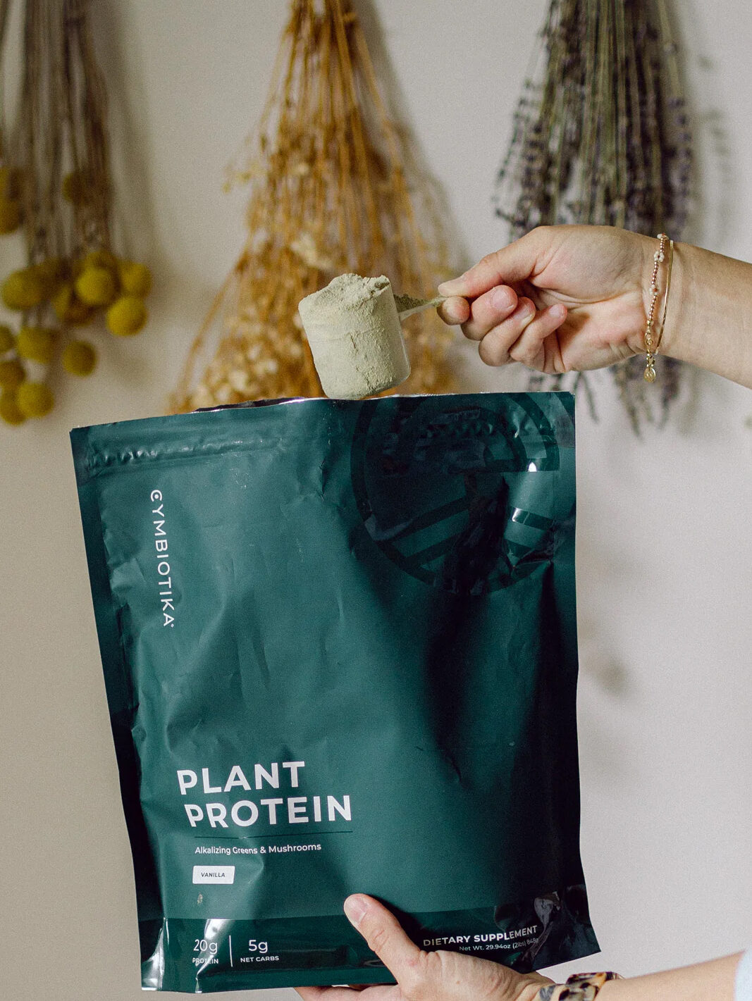 A hand scoops powder from the bag in front of a wall of drying plants