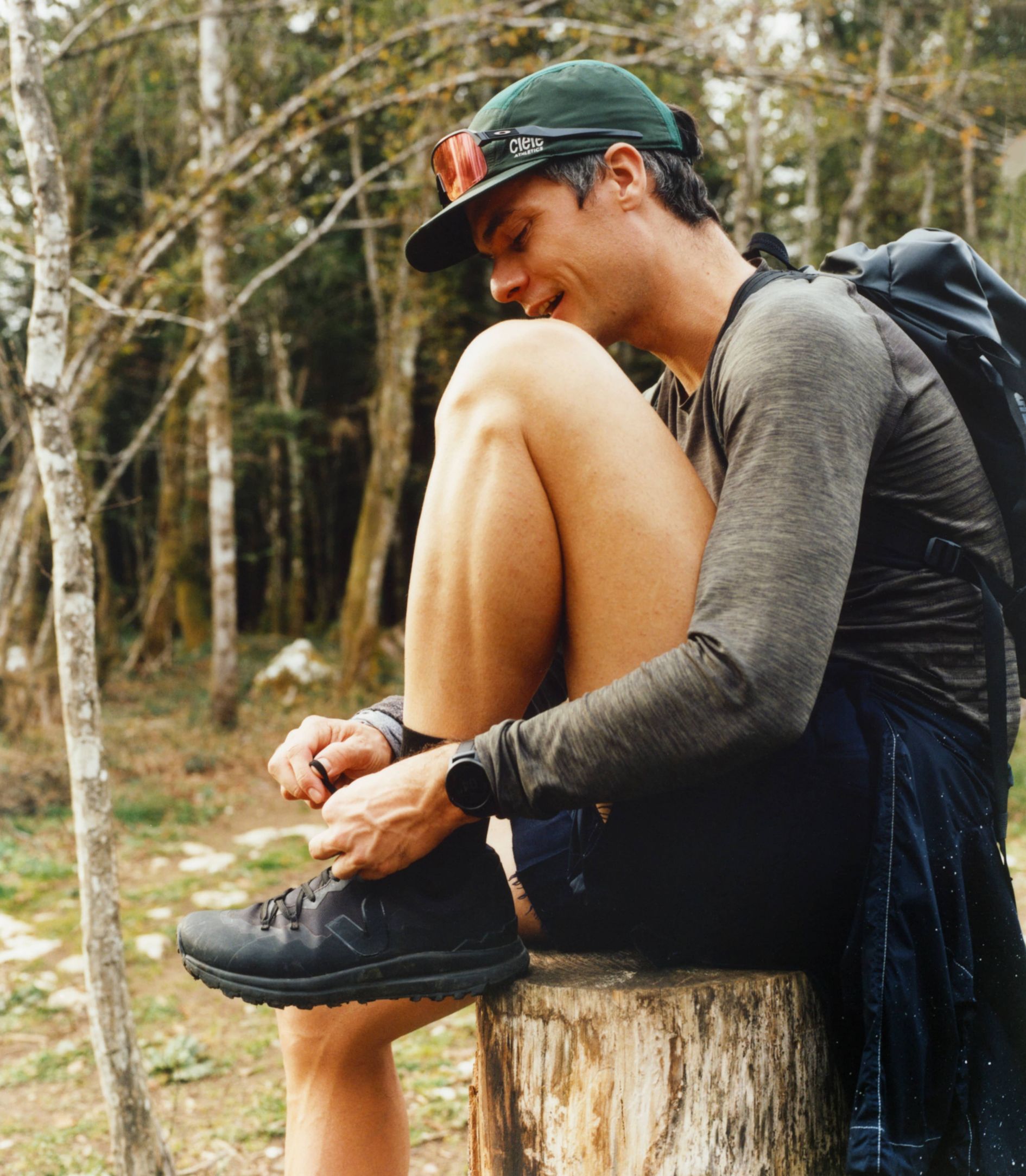 A man ties his shoes while sitting on a stump in the outdoors.