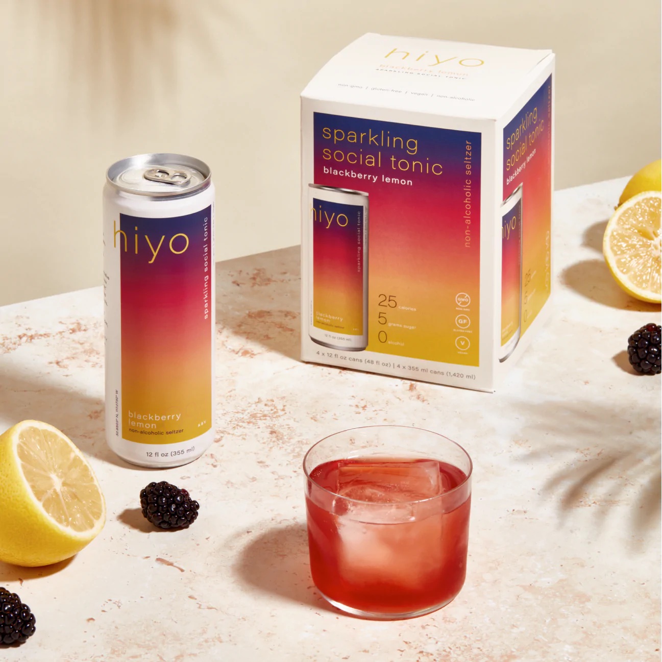 A box of hiyo blackberry lemon tonic with a can next to it, and a glass of the tonic with a large ice cube and a blackberry on the table.