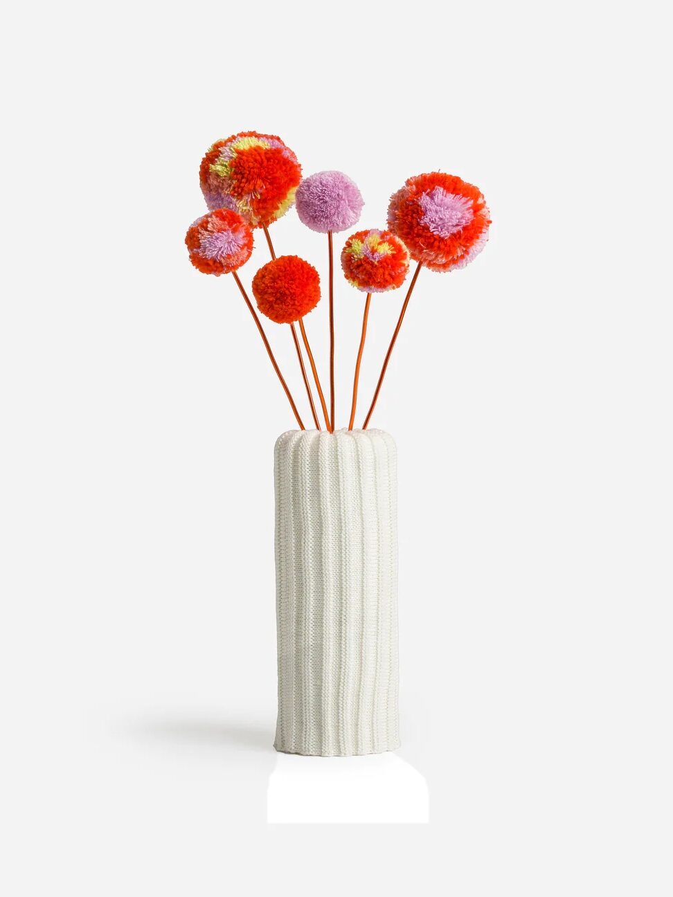 A white knit vase holds a red and lavender bouquet of poms