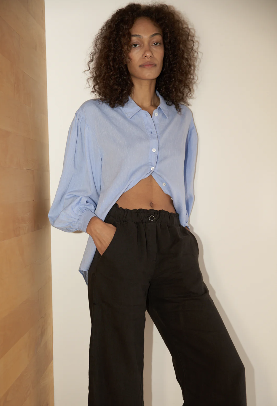 A model leans against the wall in a blue button down with her hand in the pocket of her black linen pants. 
