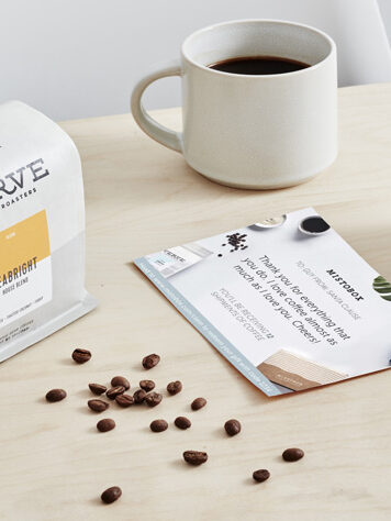 A cup of coffee, a bag of coffee, and some loose beans sit next to a postcard from the brand on a wood table top.