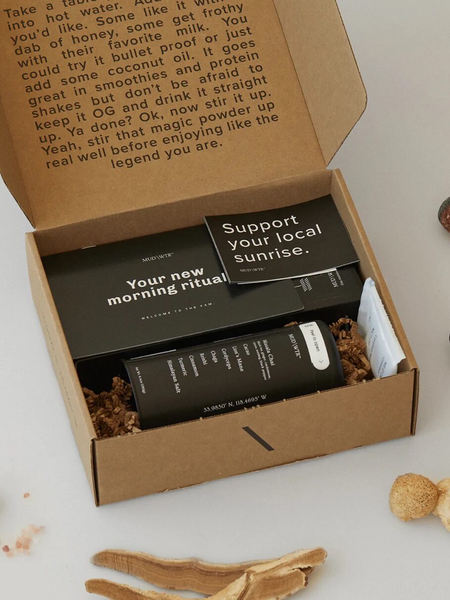A cardboard box filled with the Mud\Wtr products sits on a table surrounded by loose mushrooms