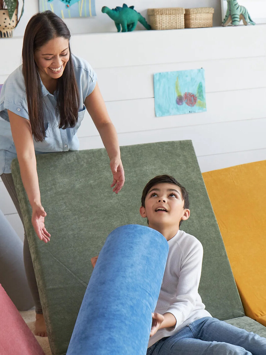 A woman leans over the back of a green play cushion and reaches for a blue cushion in a boy's arms