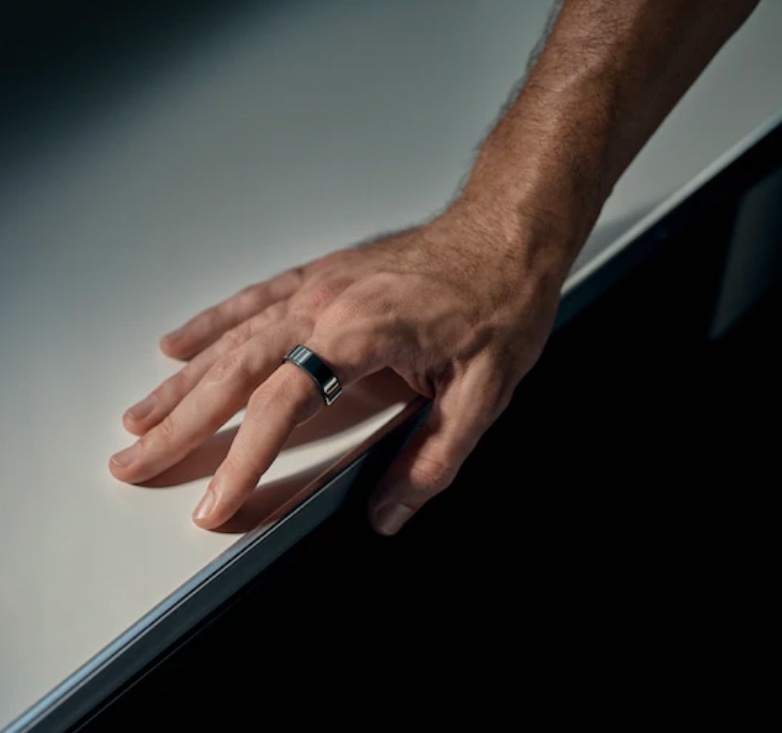 A man's hand with an Oura ring on his pointer rests on a table.