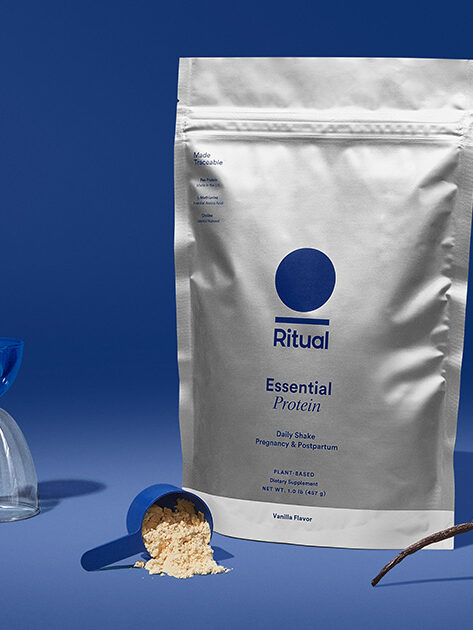 A bag of protein powder sits in a blue studio shot with a scoop of the product spilling on the surface in front of it.