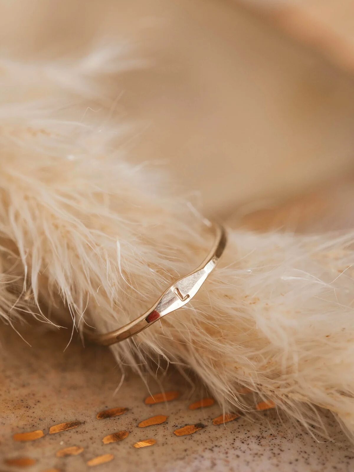 A small gold signet ring with the letter "L" sits on a faux fur background