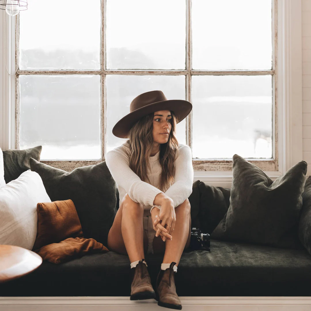 A woman sitting in front of windows wears a brown wide-brimmed hat