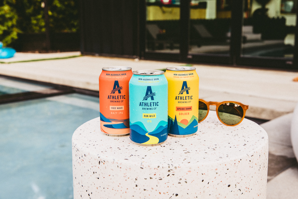 Three cans of Athletic Brewing non-alcoholic beer set on a stonetop outside by a pool.