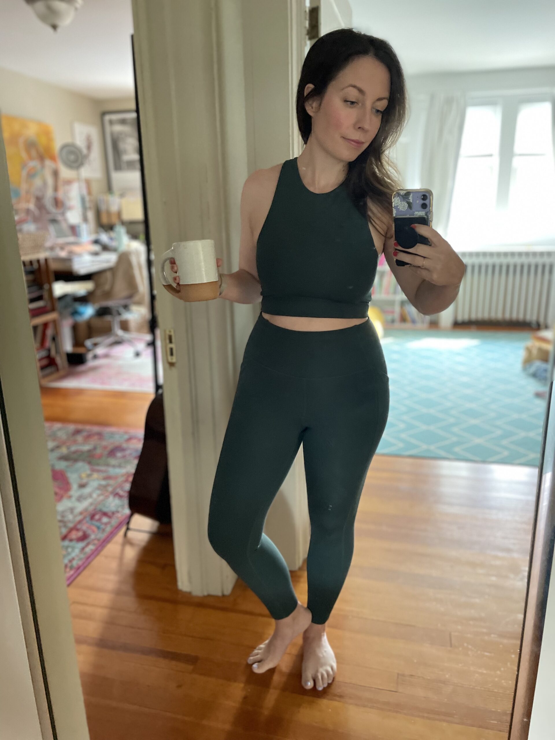 A woman takes a photo of herself wearing a sports bra and leggings in a full length mirror.