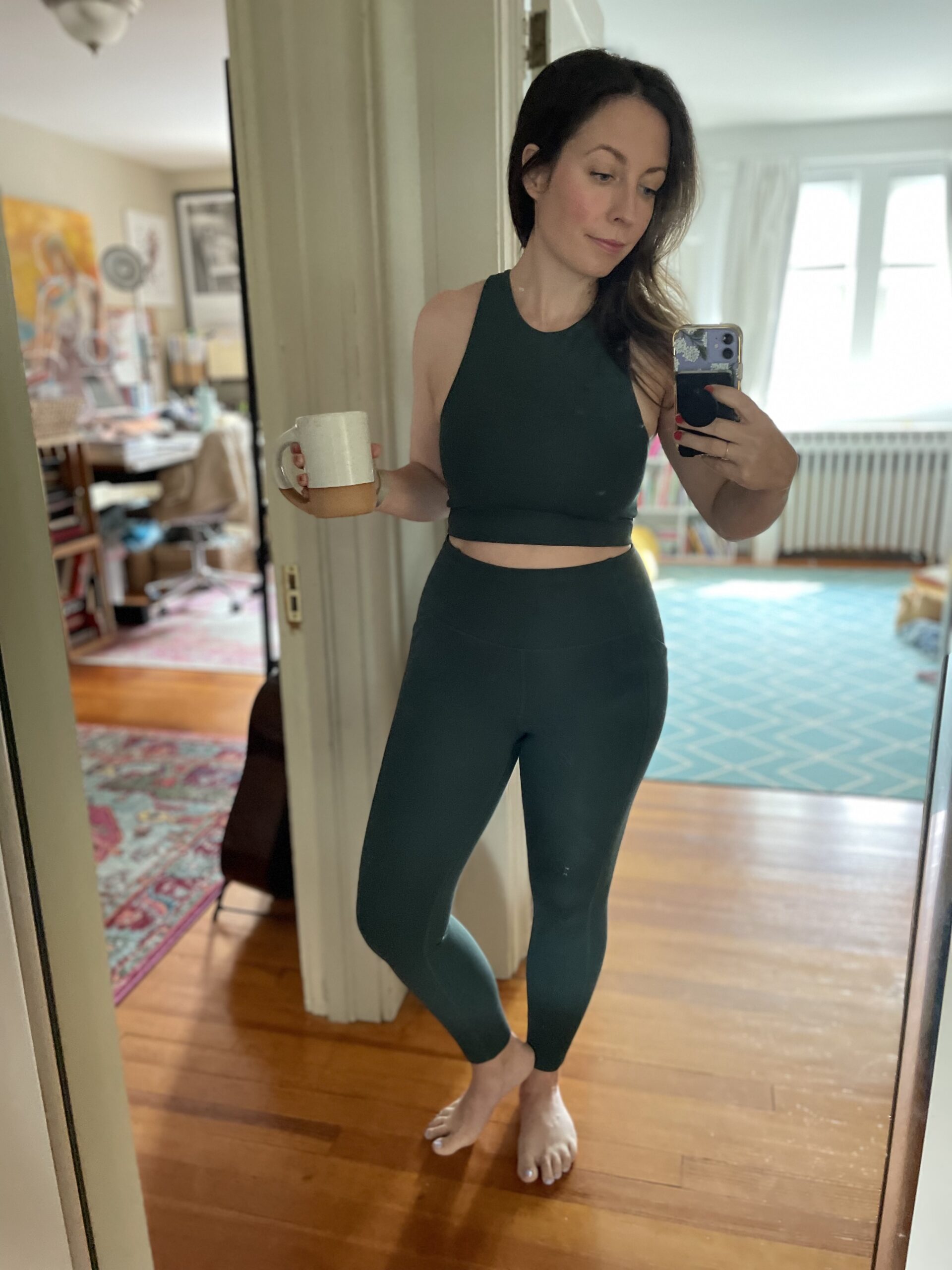 A woman takes a photo of herself wearing a sports bra and leggings in a full length mirror. 