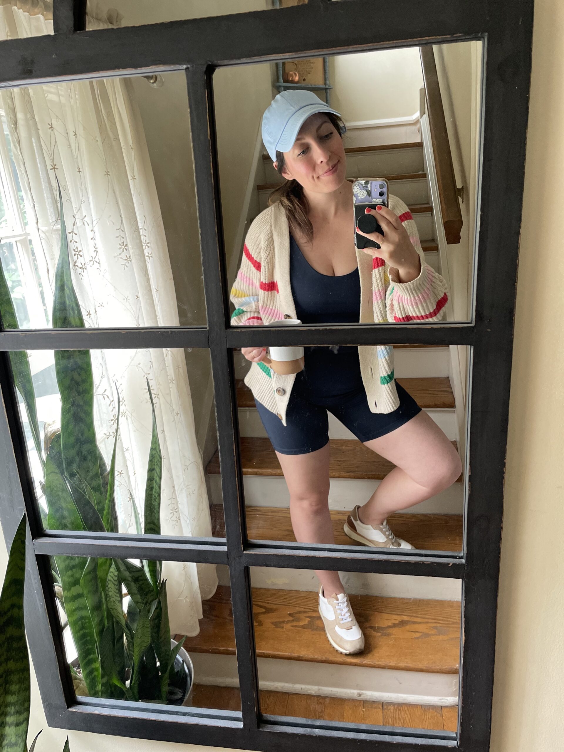 A woman holding a coffee mug takes a photo of herself in a mirror with her phone: she's wearing white and tan sneakers, a unitard, a striped cardigan, and a blue baseball hat.