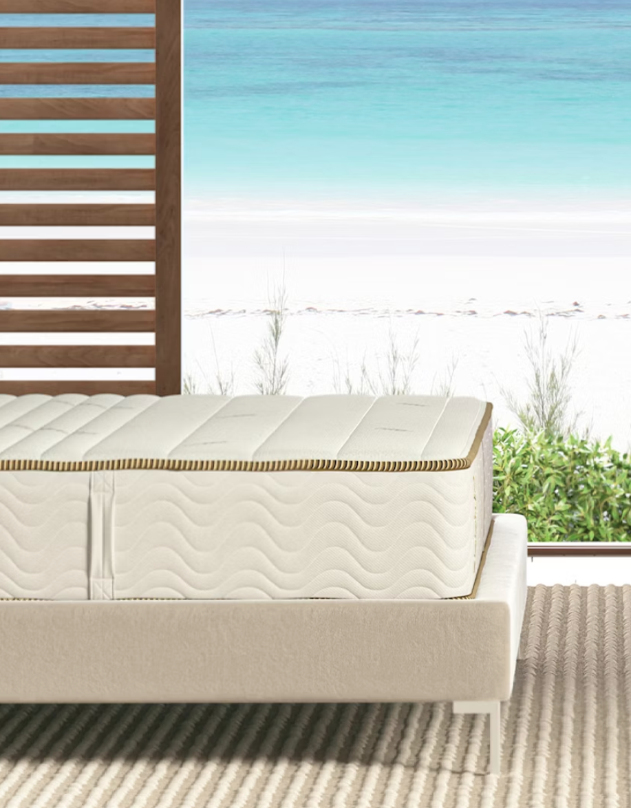 An uncovered latex mattress sits in a platform bed next to an open window overlooking the beach.