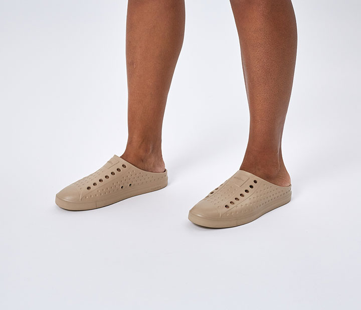 A pair of men's legs wearing tan slip on mule-style Native shoes. 