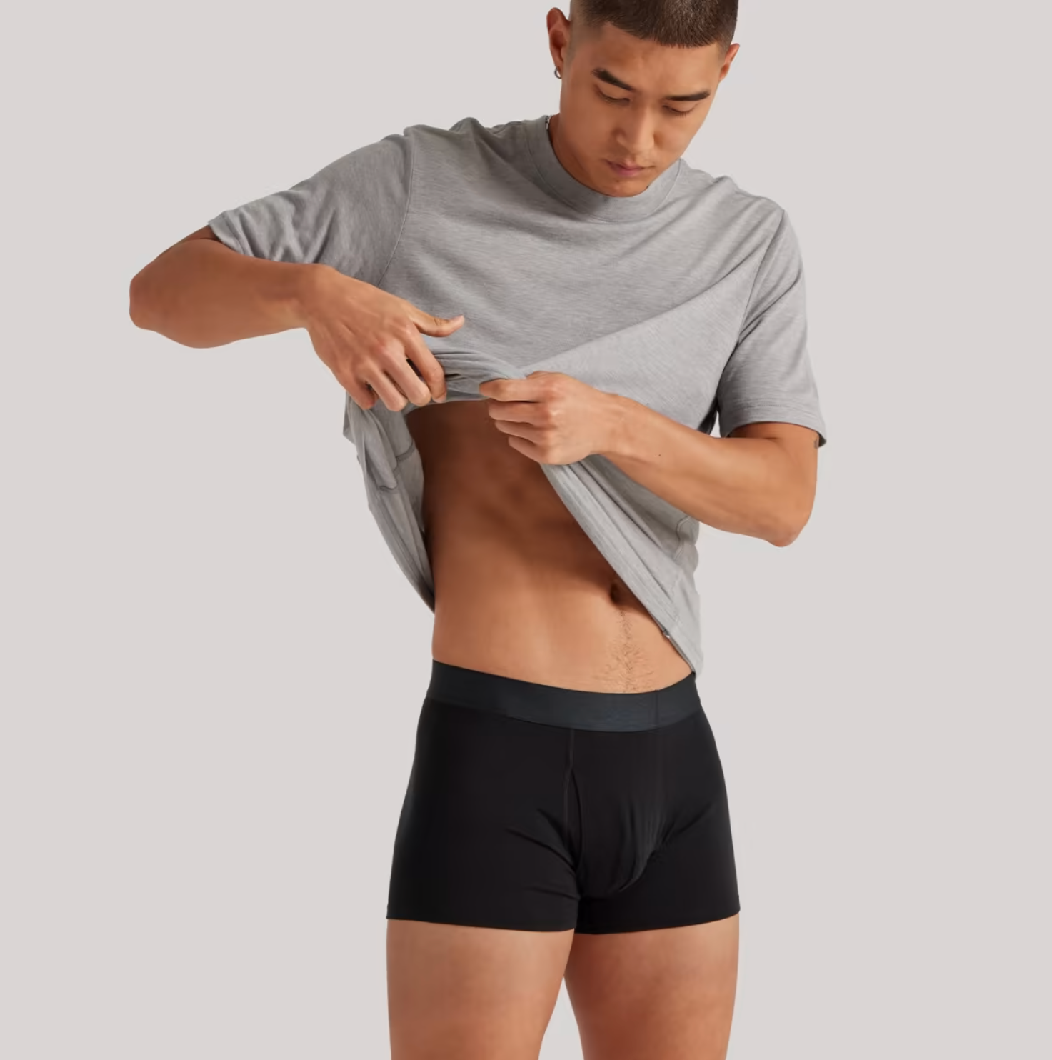 A model holds up his gray t-shirt to show off his black boxer briefs.