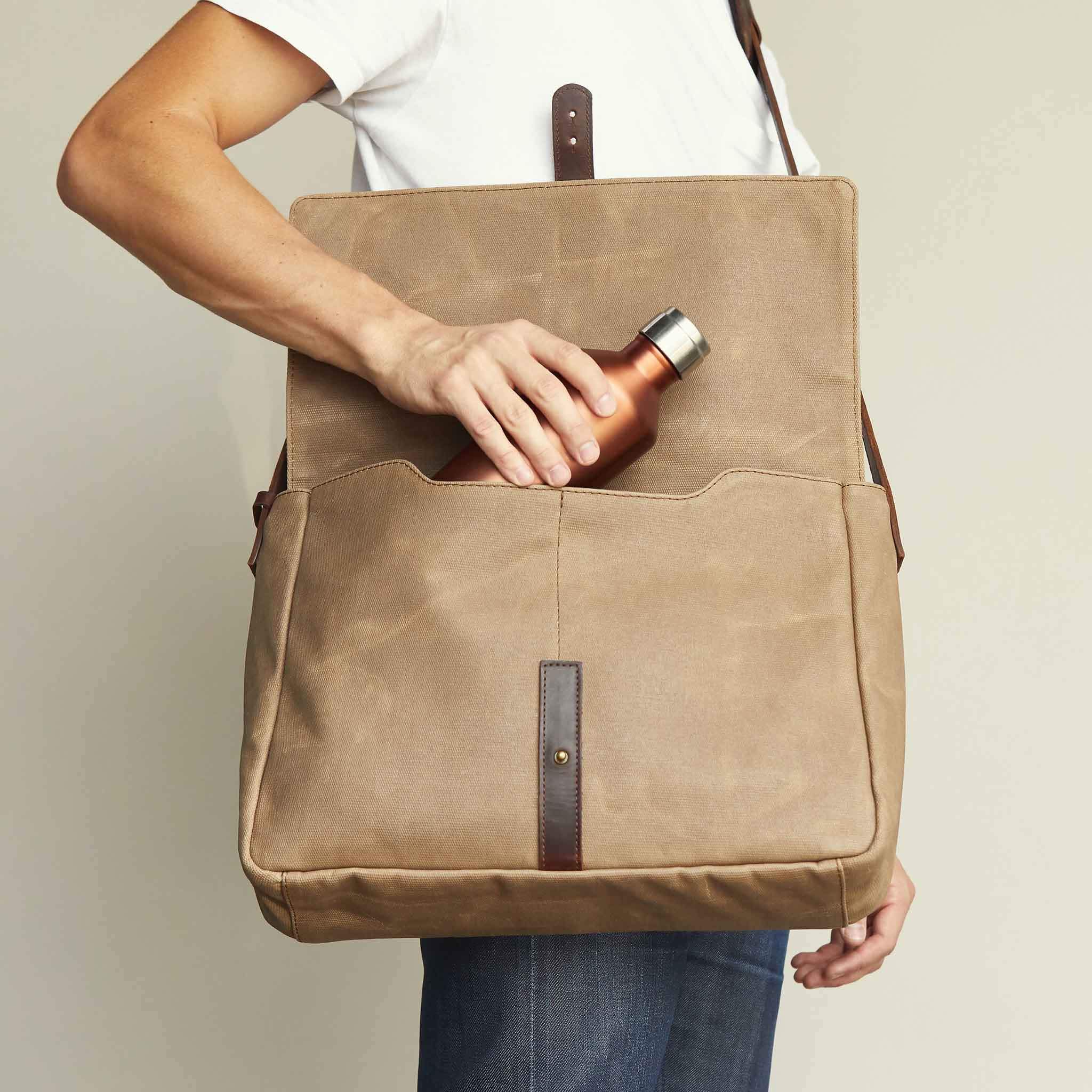 A man tucks a steel water bottle into a waxed canvas messenger bag. 