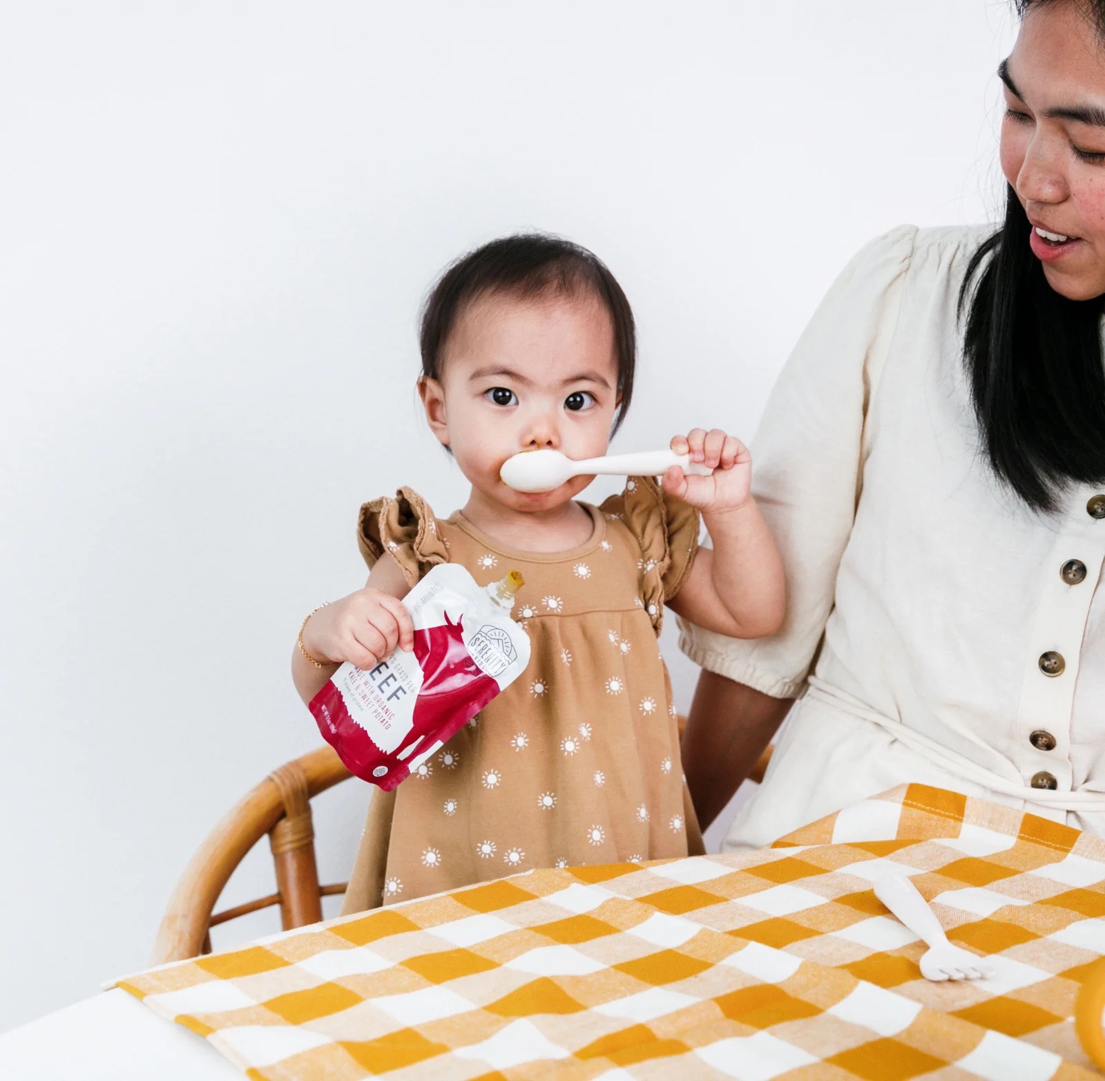 A young child stands at a table with a beef pouch and holds a spoon to her mouth. 