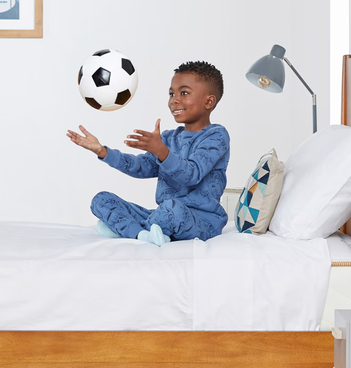 A little boy throws a soccer ball while he sits on a Saatva bed. 