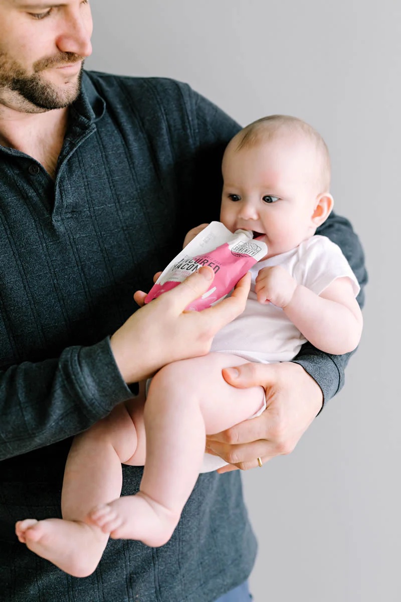 A man holds a baby and puts a bacon pouch to its mouth. 